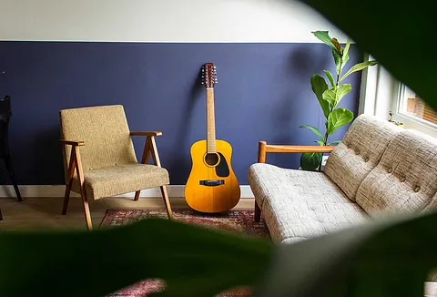 Akustikgitarre lehnt an einer lila Wand zwischen einem Vintage-Sessel und einem Sofa.