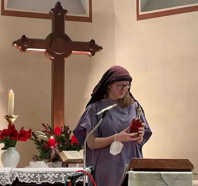 Person in einer Tunika steht vor dem Altarkreuz einer Kirche.